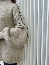 Buzo oversize de punto color beige, con cuello alto, mangas largas abullonadas y detalles de costuras decorativas bordadas a mano en color negro a lo largo de los hombros y mangas.