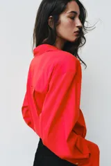 Camisa de popelina roja, de corte oversize, con cuello solapa, manga larga y bajo asimétrico redondeado. Cierre frontal oculto con botones.