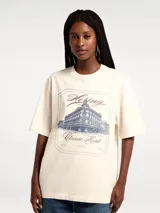 Camiseta de corte oversized color beige, con cuello redondo y mangas cortas. Presenta un estampado frontal estilo vintage con la ilustración de un edificio y la inscripción 'Hering Classic Hotel'.