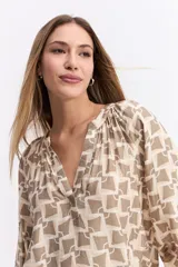 Blusa de corte relajado con estampado geométrico en tonos beige y negro. Presenta cuello en V con detalle de frunces, mangas largas abullonadas con puños elásticos y una caída fluida.