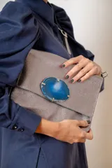 Sobre de cuero color suela con solapa frontal decorada con una piedra de ágata natural enmarcada en metal. Incluye correa larga ajustable y extraíble para usar como bandolera.