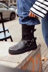 Bota negra de cuero con caña media, suela track, correa con hebilla en el tobillo y arnés con argolla metálica.