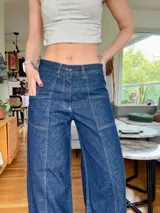 Jean de tiro medio en color azul índigo oscuro, con corte de pierna ancha y costuras verticales decorativas en el frente. Presenta bolsillos delanteros tipo parche y cierre de botón y cremallera.