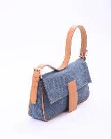 Cartera tipo baguette confeccionada en tela de jean azul con detalles en cuero color suela. Presenta una correa ajustable con hebillas metálicas, solapa frontal con cierre magnético y un aplique de cuero con logo grabado.