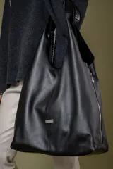 Bolso tipo tote de gamuza en color verde oliva, con diseño minimalista y asa integrada. Presenta una placa metálica con el logo de la marca en la parte inferior central y detalles de textura en el asa.