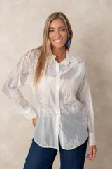 Camisa de manga larga en color blanco, confeccionada en tejido traslúcido con delicados bordados florales en hilo de seda al tono. Presenta cuello clásico, cierre frontal con botones y puños abotonados.