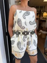 Conjunto de dos piezas (top y short) de satén color crema con estampado paisley en negro y detalle de borde estampado en tonos dorados/mostaza. El top es de tirantes finos y el short es de tiro alto con elástico y cinturón de cuero negro con tachas.