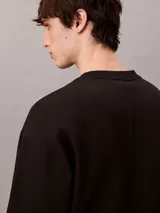 Buzo Calvin Klein de felpa color negro, con cuello redondo, manga larga, puños y dobladillo acanalados. Presenta un bordado frontal con la palabra "VERY" en tonos verdosos y debajo el logo de la marca.