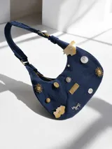 Cartera tipo baguette de gamuza azul marino con diseño decorado con múltiples tachas, botones y dijes metálicos de diversas formas. Cuenta con una correa de hombro integrada y placa metálica con logo de la marca en la parte frontal.