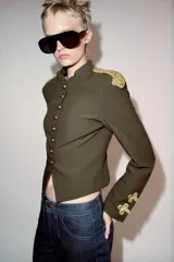 Cazadora militar de color verde oscuro, de cuello subido y manga larga. Cuenta con detalles de alamares y cordones dorados en los hombros y puños, y cierre frontal con botones dorados metálicos.