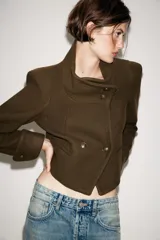Chaqueta corta de paño color verde militar, con cuello subido, manga larga con puños abotonados y cierre frontal cruzado con botones metálicos. Presenta hombreras y bolsillos de vivo en el delantero.