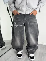 Pantalón de jean estilo baggy con lavado desgastado en color gris oscuro. Presenta bordado con la frase 'Stay real' en el muslo derecho y 'different' en la parte inferior de la pierna izquierda.