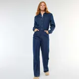 Mono de jean azul oscuro con cierre frontal de botones, cuello camisero, mangas largas y bolsillos frontales. Presenta un corte recto y holgado en las piernas.