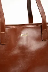 Bolso tipo tote bag color marrón, confeccionado en cuero sintético, con doble asa superior y detalle de placa metálica dorada con logo en la parte frontal.