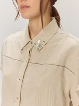 Camisa de manga corta color beige con sutil diseño a rayas verticales, cuello camisero con detalle de botones decorativos en la solapa y cierre frontal oculto.