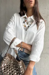Camisa blanca de corte clásico con cuello camisero y cierre frontal de botones. Presenta un delicado bordado tono sobre tono en las mangas y una textura sutil en la tela.