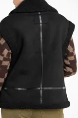 Chaleco estilo gamulán de color negro, con interior y detalles de corderito sintético. Presenta cierre frontal, cuello solapa y bolsillos laterales.