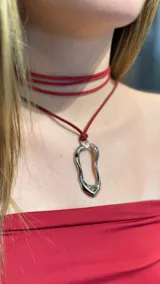 Gargantilla tipo choker de gamuza roja con diseño de doble vuelta en el cuello y dije colgante de metal plateado con forma orgánica irregular.