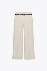 Pantalón de vestir de tiro alto, color beige claro, de corte ancho y pierna recta. Presenta pliegues marcados en el frente y puños anchos doblados en el bajo. Incluye un cinturón fino negro con hebilla metálica.