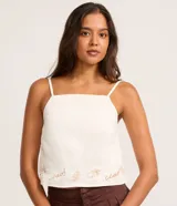 Blusa confeccionada en sarga de algodón, de color blanco, con escote cuadrado y tirantes finos. Presenta un detalle bordado en la parte inferior con motivos botánicos y la palabra ciao.