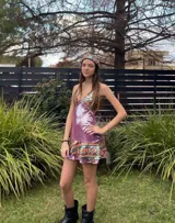 Vestido corto de tela metalizada en tono fucsia con reflejos dorados, con escote en V, diseño de top con triángulos, aberturas laterales en la cintura y doble volado irregular en el ruedo.