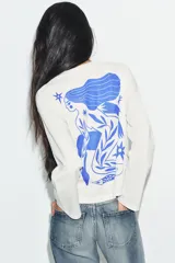 Remera de manga larga y corte oversize, color blanco, con cuello redondo. Presenta un estampado gráfico en color azul en el pecho y a lo largo de la manga derecha.