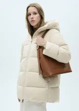 Campera acolchada tipo puffer de color beige, con diseño recto, cuello alto y capucha integrada. Presenta cierre frontal de cremallera doble y bolsillos laterales con cierre.