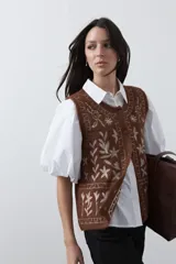 Chaleco tejido color marrón con bordados artesanales de motivos florales y geométricos en color crema. Presenta cuello redondo, cierre frontal con botones y diseño sin mangas.