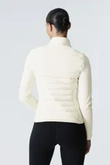 Campera híbrida color crema, con cuerpo acolchado y mangas de tejido elástico. Presenta cuello alto, cierre frontal y bolsillos laterales con cierre.