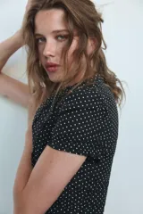 Camisa negra con estampado de lunares blancos, cuello redondo con escote en pico, mangas cortas abullonadas y detalle de volantes frontales. Cierre frontal con botones.