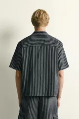 Camisa de corte relajado confeccionada en denim de algodón y lino, con diseño de rayas verticales, cuello solapa y manga corta con cierre frontal de botones.