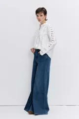 Camisa de algodón color blanco con detalles de encaje broderie y alforzas verticales. Presenta cuello alto con volados, cierre frontal de botones y mangas largas con puños acampanados.