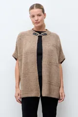 Capa tejida de punto en color beige, con diseño abierto y cierre frontal mediante broche metálico con detalles en cuero sintético negro. Presenta mangas cortas y textura suave.