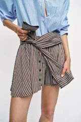 Bermuda de tiro medio con estampado de rayas verticales en tonos marrones y celestes. Presenta un diseño único con lazada frontal que simula mangas anudadas, elástico en la parte posterior de la cintura, cierre frontal con cremallera y botones, y bajo con vuelo.