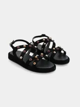 Sandalias planas de cuero gamuza en color negro, con múltiples tiras cruzadas decoradas con tachas metálicas doradas y aplicaciones de piedras rojizas. Cuenta con suela de PVC y tira ajustable en el talón con hebilla metálica dorada.