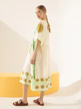 Túnica midi de color blanco crudo, con mangas cortas amplias, escote en V con cordones verdes ajustables y cintura fruncida. Presenta detalles de bordados geométricos verdes y soles marrones en las mangas y el ruedo.
