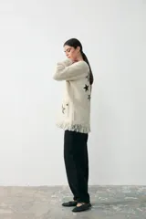 Chaqueta abierta de punto color crema, con diseño de corte recto, bolsillos frontales, flecos en el ruedo y detalle de estrella bordada en uno de los bolsillos.