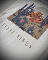 Hoodie oversize color crema con estampado en la espalda que representa un paisaje desértico con cactus y montañas, acompañado por el texto 'DESERT VIBES'.