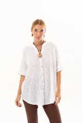 Camisa de broderie blanca con silueta oversize, escote en V, cierre frontal con botones y mangas 3/4. Presenta un diseño con bajo asimétrico, siendo ligeramente más larga en la parte posterior.