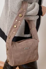 Cartera de hombro confeccionada en gamuza color marrón, con diseño de asa ancha decorada con tachas metálicas doradas de diversos tamaños. Presenta cierre superior y placa metálica con el logo de la marca en el frente.