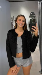 Chaqueta corta de textura peluda en color negro, con cuello redondo y cierre frontal abierto. Presenta mangas largas y un corte recto.