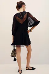 Vestido corto negro con transparencias, mangas largas amplias y detalles de bordados florales con lentejuelas en el frente. Cuenta con un lazo ajustable en la cintura y falda fluida.