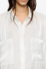 Camisa oversize de manga larga, color blanco, confeccionada en viscosa brillante. Presenta cuello camisero, cierre frontal de botones y dos bolsillos de parche en el pecho.
