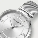 Reloj analógico Mini Focus con caja redonda de metal plateado y malla de acero tipo milanesa. Presenta una esfera minimalista con índices de bastón y números romanos en las posiciones 3, 6, 9 y 12.