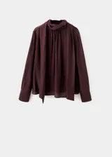 Blusa de cuello alto con detalle de fular integrado, confeccionada en tejido con textura. Presenta diseño recto, manga larga con puños abotonados y cierre posterior.