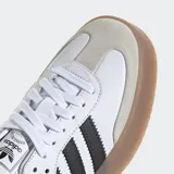 Zapatillas Adidas Sambae blancas con las tres tiras laterales en negro, refuerzo de gamuza gris en la puntera, contrafuerte trasero negro y suela de goma color caramelo.