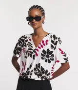 Blusa de viscosa con escote en V y mangas cortas abullonadas. Presenta un estampado de follaje y flores en tonos negro, fucsia y beige sobre fondo blanco.