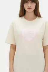 Remera de algodón color beige con estampado frontal de un corazón rosado que contiene un moño. Tiene cuello redondo y mangas cortas.