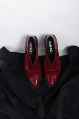 Mocasines de charol color bordó, de diseño vanguardista con corte asimétrico en el empeine, abertura en V, punta fina y suela plana con pequeño tacón.