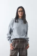 Polera de tejido de punto color gris, con cuello alto, mangas largas abullonadas con puños elásticos y costura central vertical en el frente. Presenta una abertura sutil en la espalda.